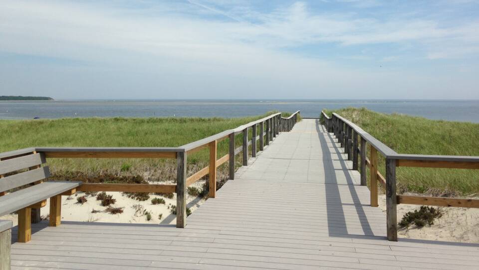 A raised wooden boardwalk leads out, over marsh grass and sand, toward a wide body of water.