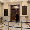 A court officer in a blue uniform opens the door to the House chamber inside the Massachusetts State House, with a stack of papers in his hand.