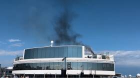 Black smoke rises above a large cruise ship and into the blue sky.