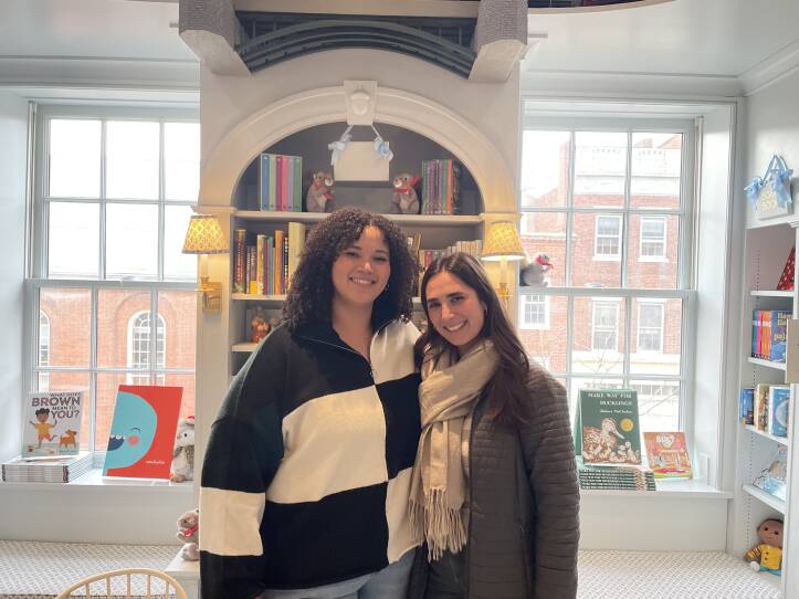 Two young women stand next to each other inside a bookstore. One is wearing a black and white checkerboard patterned top. Her friend stands to her right. She has a large, beige scarf wrapped around her neck and a light grey winter coat, which is unzipped. They're standing in front of a bookcase and reading nook. It appears to be a room with children's literature as toys, stuffed animals and a copy of "Make Way for Ducklings" are all on display.