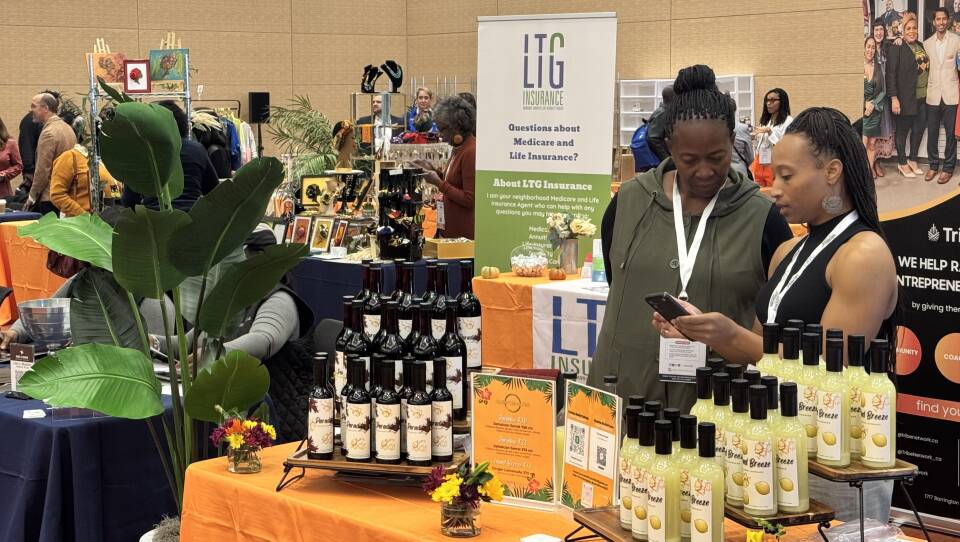 Two women in the foreground talking behind a table with bottles and products on it.