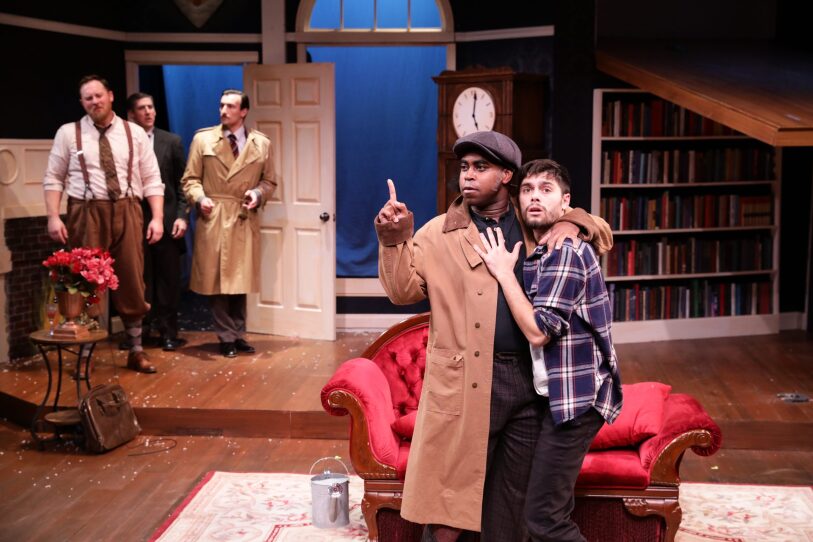 This is a scene from The Play That Goes Wrong. In this image two men cling to each other,.they appear to be frightened. In the background three men have entered the apartment. The mood is one of chaos.