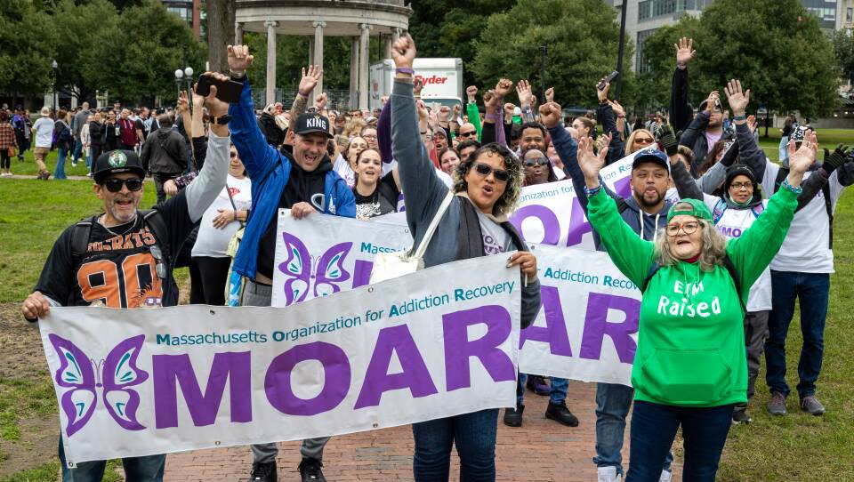 A large crowd of people in a park hold their hands up and cheer. A few people in the front are holding signs for "Massachusetts Organization for Addiction Recovery"