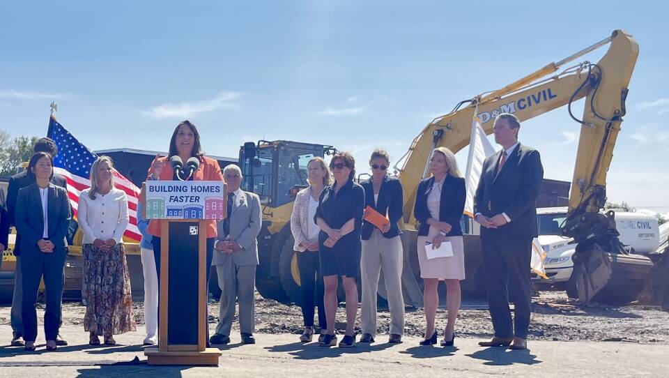 A woman in an orange blazer stands at a wooden lectern at a construction site, with several other people and yellow construction vehicles behind her.