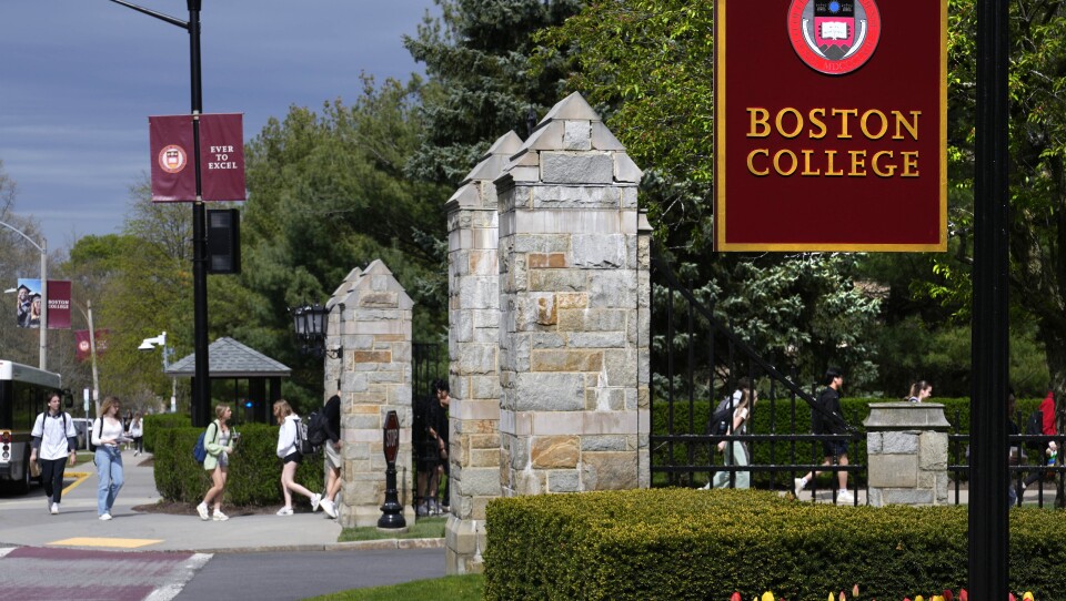 About a dozen students wearing backpacks walk along the sidewalk at the entrance to Boston College.