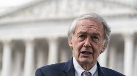An older man wearing a suit jacket and tie stands in front of the Supreme Court of the United States.