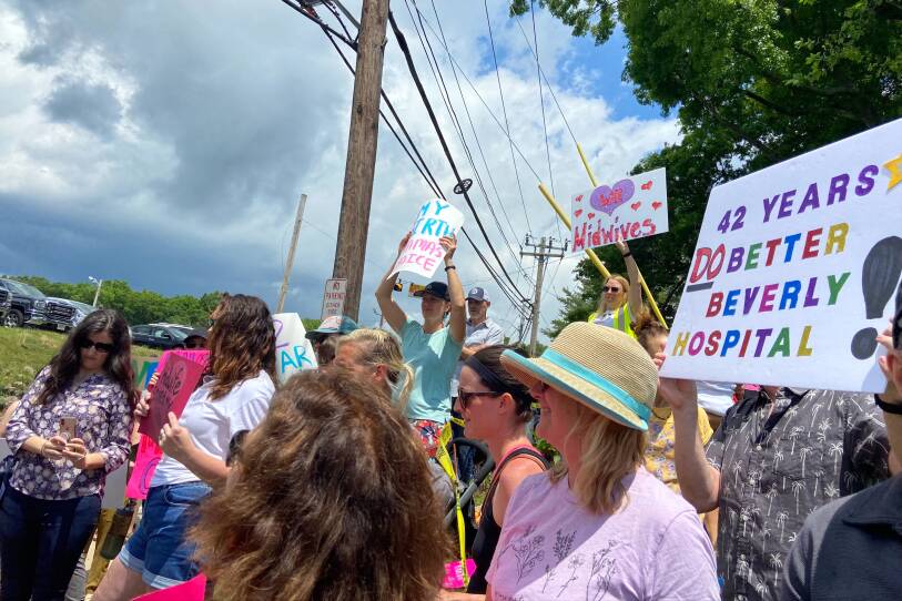 A group stand with signs on a sunny day, with signs reading "We Love Midwives" and "42 years: Do better Beverly Hospital"