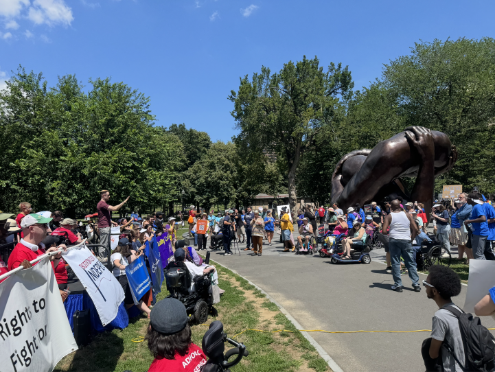 Hundreds of people stand on the Common in front of the bronze Embrace statue. Many hold up signs and some are in wheelchairs.