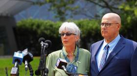 Standing behind a podium with several news microphones are: A woman with gray hair in a blouse and thin sunglasses beside a bald man wearing thin, dark-rimmed glasses and a suit and tie. They're looking somberly over the podium.