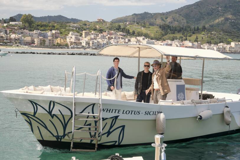 Adam DiMarco, Michael Imperioli, and Murray F Abraham stand on a boat with the words "White Lotus Sicily" written on the side of it in a river in Italy