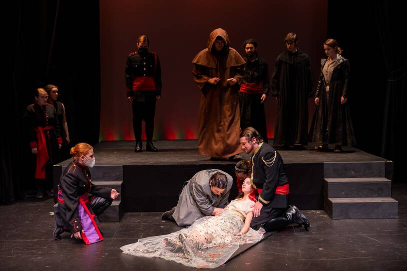 On stage, a woman lies on the ground as people attend to her. They are surrounded by people wearing Shakespeare costumes.