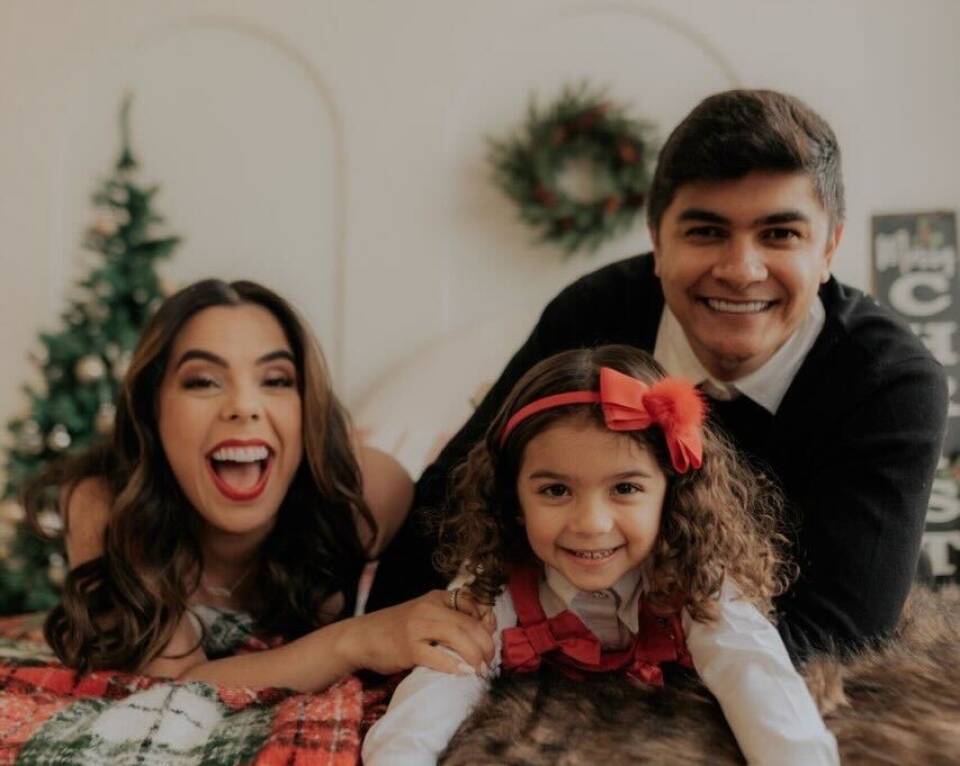 A happy family with two parents and a young girl smile as they lean over the back of a couch in their living room.