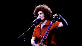 Female musician dressed in red jumpsuit tunes her guitar during a performance.