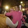 A smiling woman in a pink wool coat is on a city street at night, flanked by two other women and a crowd holding campaign signs.