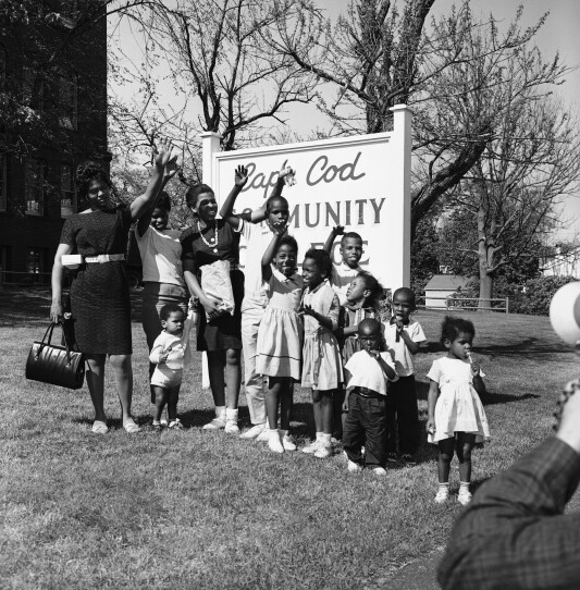 A Black women stands with 11 children, as they all smile and wave in front of a Cape Cod Community College sign