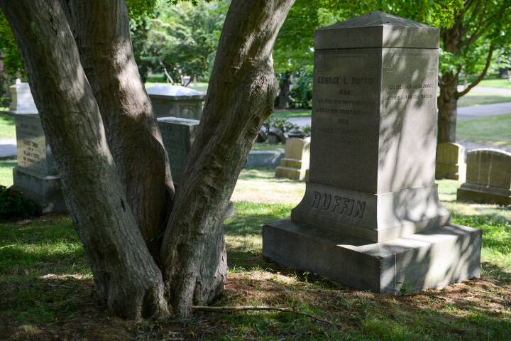 A grey marble monument stands in a cemetery beside a tree that branches off in three directions. The monument reads RUFFIN in large letters at the base, and higher up, George L Ruffin and Josephine St. Pierre Ruffin.