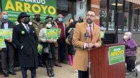 A man in a tan jacket and tie speaks into a microphone at a podium in the street. A small group of people holding "Richard Arroyo" campaign signs are behind him on the sidewalk.