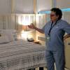 A woman in blue medical scrubs gestures with one hand toward a freshly made bed in a large, dimly lit room.