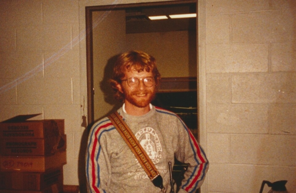 This is a photograph of Brian O'Donovan in 1984. We see him from the waist up. He appears to be in a break room of a building as there is a coffee pot and boxes in the background and fluorescent lights above. He has a mop of read hair. He's looking directly at the camera and smiling.