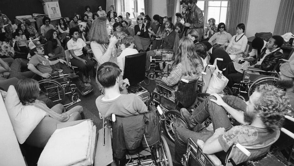 A black and white photo. A large group of people, mostly in wheelchairs gatheraround a woman in the center who is signing.