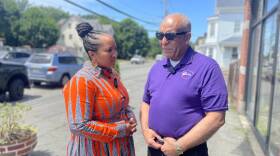 A woman in a dress stands facing a man in a purple shirt and sunglasses on a sidewalk next to a road and a building with windows.
