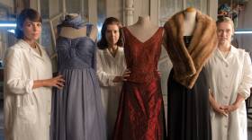 Three women in long white coats standing with couture dresses on mannequins