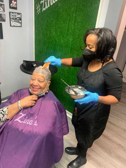 A hair stylist stands at her client and applies a treatment. The client wears a purple robe.