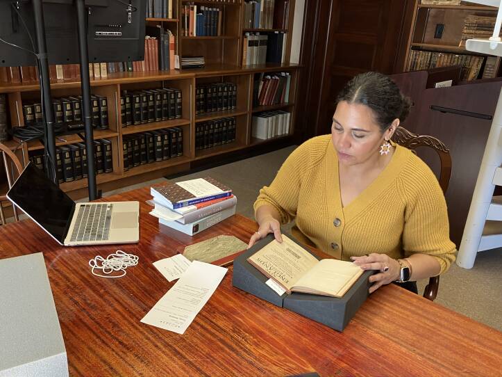 A woman sits in an office surrounded by book cases. She looks down at an old book.