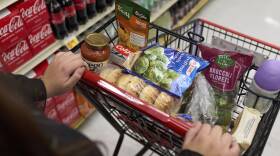 A person pushes a small shopping cart through a grocery store aisle. It only has a handful of items loaded, including bagels, broccoli, toothpaste and pasta sauce.