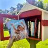 A small, wooden box painted red holds books open to anyone. Photo shows someone holding up a book in front of the collection of books in the library called "Juneteenth for Mazie" by Floyd Cooper.