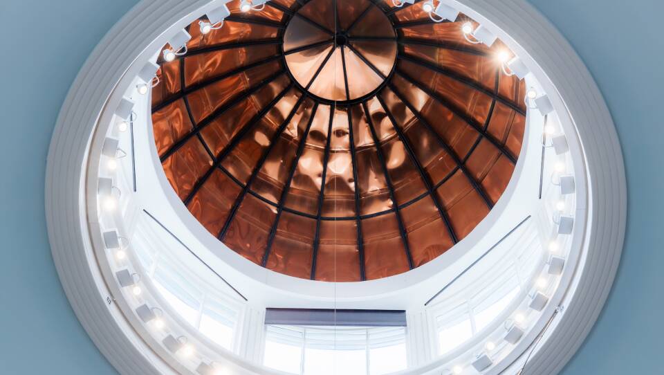 A large, circular cupola is rimmed with shiny copper atop bright, white windows and a row of lights encircling the dome.