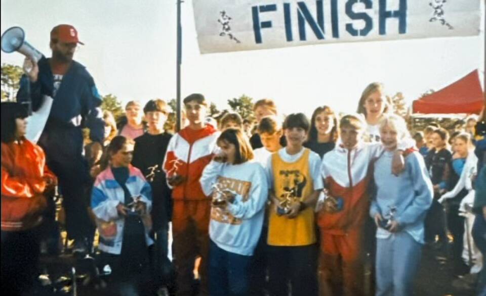 Dozens of children crowd around a finish line on a field, with a row of girls holding trophies in the front.
