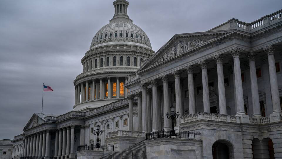 The U.S. Capitol is shown against a grey evening sky, with glowing amber light visible through the windows of the rotunda.