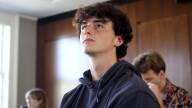 A teenage boy with fluffy brown hair sits in an academic room, other students behind him. He seems to be listening attentively.