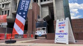 A fabric pop-up banner in front of a large government building says "vote here". A second sandwich board says "early voting here".