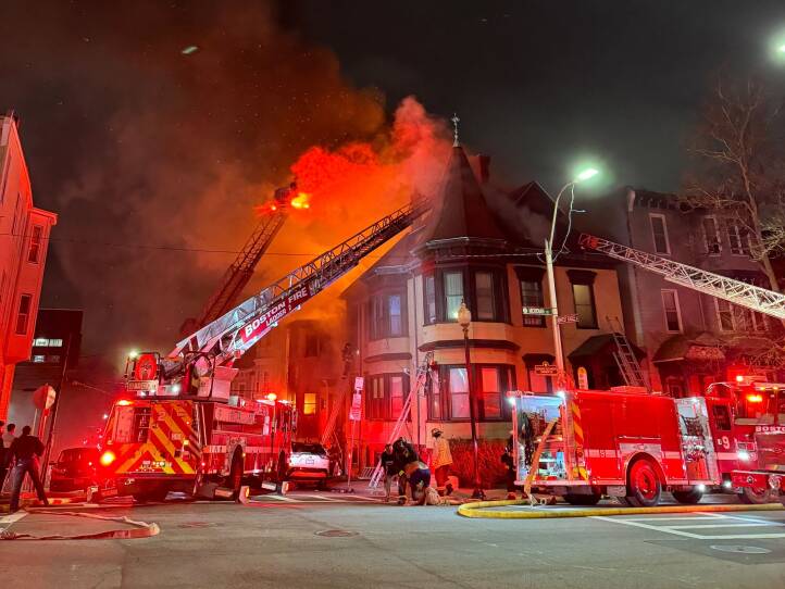Red lights of multiple fire trucks light up a residential building and plume of smoke.