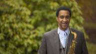 Javon Jackson wears a grey and blue suit and holds a tenor saxophone across his chest.