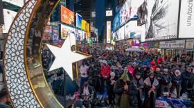 A large crescent moon and star made of metal overlooking a large group of people in a city street, standing with their heads bowed in prayer.