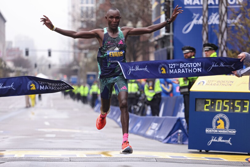Chebet runs through the tape at the Boston Marathon finish line, his hands out to his side. It's a gray cloudy day.