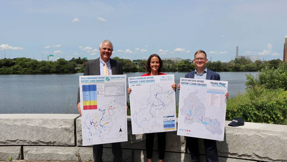 Three people, standing side-by-side, hold up posters with illustrated images of the Neponset, Charles and Mystic rivers as the trio stands on a lookout by the edge of a river.