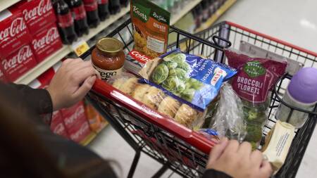 A person pushes a small shopping cart through a grocery store aisle. It only has a handful of items loaded, including bagels, broccoli, toothpaste and pasta sauce.