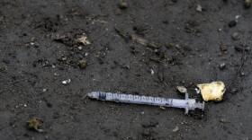A syringe with a bent needle is shoved into the dirt ground.
