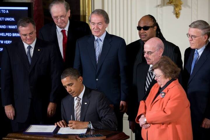 At an indoor ceremony, President Barack Obama sits at a small wooden desk, pen in hand, signing a stack of papers.