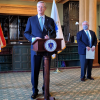 A man stands at a lectern in the middle of the room, with bookcases, a statue bust and an American flag in the background, with Lieutenant Governor Karyn Polito standing behind him.