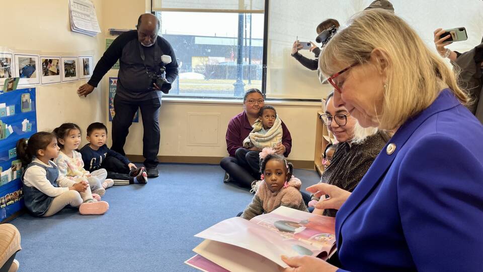 Children sit on the blue carpeted floor of a day care center, while a blond-haired woman with red glasses and a blue blazer reads a picture book to them.