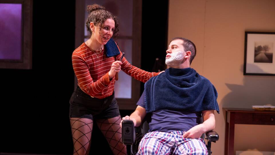 A man with shaving cream on his face sits in a chair with a towel around his shoulders. A woman with a razor stands next to him as they talk.