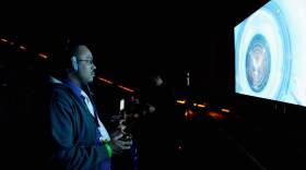 A man with large headphones stands in a dark room, playing a video game.
