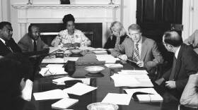 Six people sit around a large table in the White House, listening intently to President Jimmy Carter.