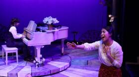 A woman sings onstage during "Preludes" at Lyric Stage Company. Behind her, a man plays piano and another lies on the ground.