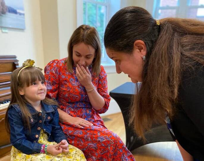 A young girl sits beside her guardian, looking up with some apprehension or timidity at a woman who's stooping down to talk to her.